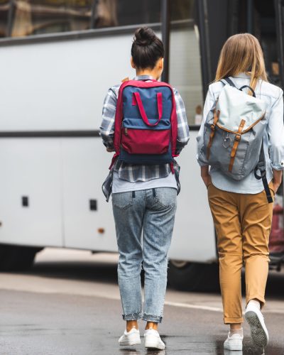 rear view of female tourists with backpacks walking near travel bus at urban street